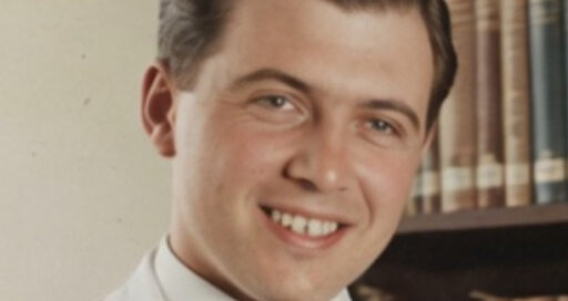 A young man with short dark hair, wearing a white coat and dark tie, smiles in front of a bookshelf filled with books.