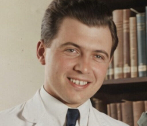 A young man with short dark hair, wearing a white coat and dark tie, smiles in front of a bookshelf filled with books.