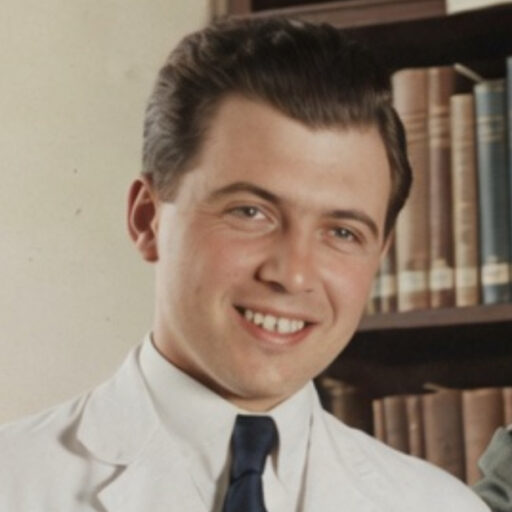 A young man with short dark hair, wearing a white coat and dark tie, smiles in front of a bookshelf filled with books.
